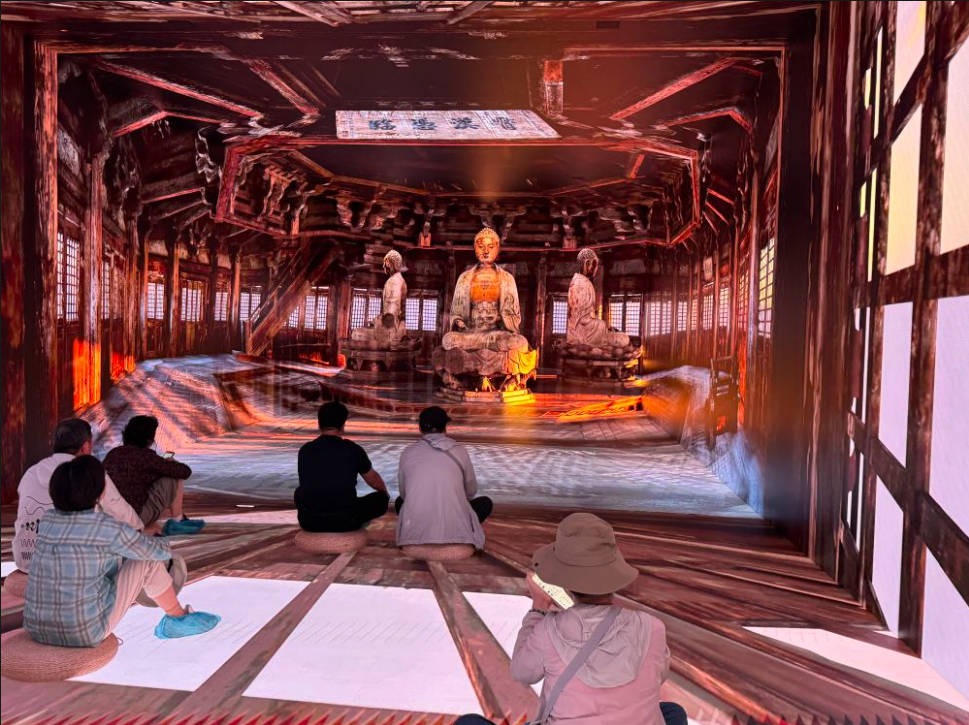The audience sits on the floor in the immersive exhibition hall, where a projection in front recreates the main Buddha statue and accompanying figures from inside the Buddhist hall of the Yingxian Wooden Pagoda; light and shadow outline the wooden beams, framework and details of the Buddha statues, showcasing the achievements of the digital conservation of cultural relics.