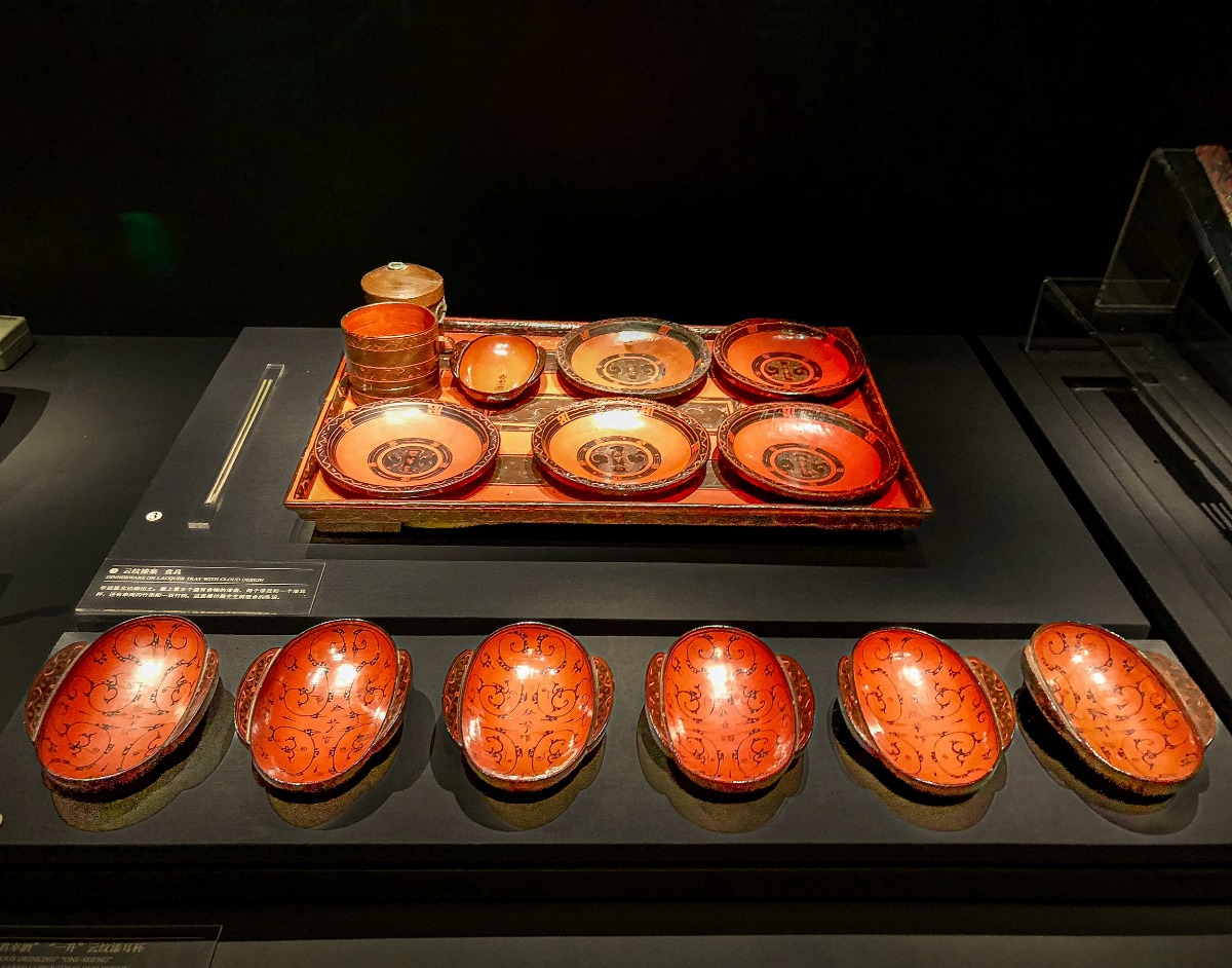 湖南省博物館展櫃中的馬王堆漢墓漆器食具：一張雲紋漆案上陳列多件紅黑漆食盤、耳杯及圓盒等器物；畫面前方一列橢圓形朱漆耳杯，杯內可見雲紋及「君幸酒」等朱書。