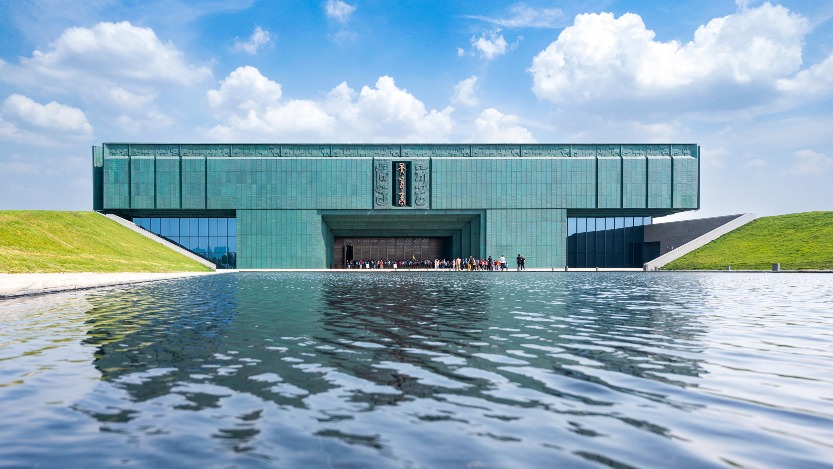 殷墟博物館新館正面外觀，建築以青銅方鼎為設計意象，大門上方寫着甲骨文「大邑商」，兩側為綠色草坡，前方水池映出館體倒影，入口處有遊客排隊。