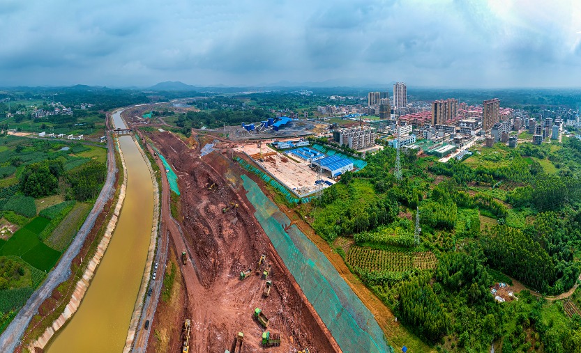 An aerial view of the Pinglu Canal construction site in Luwu Town, Lingshan County, Qinzhou City, Guangxi, with large piles of reddish-brown earth and rock next to the excavated canal channel. Multiple construction vehicles are at work on the site, with towns and farmland in the distance.