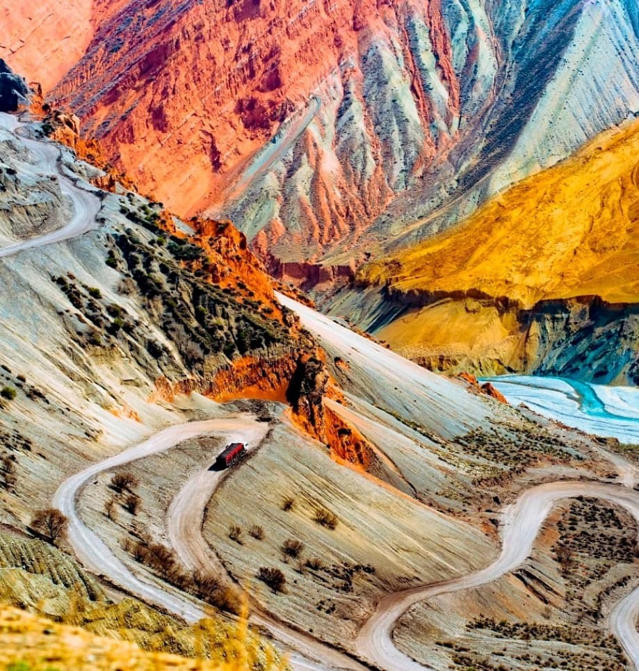 An aerial view of the Anjihai Grand Canyon in Xinjiang, where a red car drives along the road at the bottom of the canyon, contrasting with the colourful cliffs on both sides.