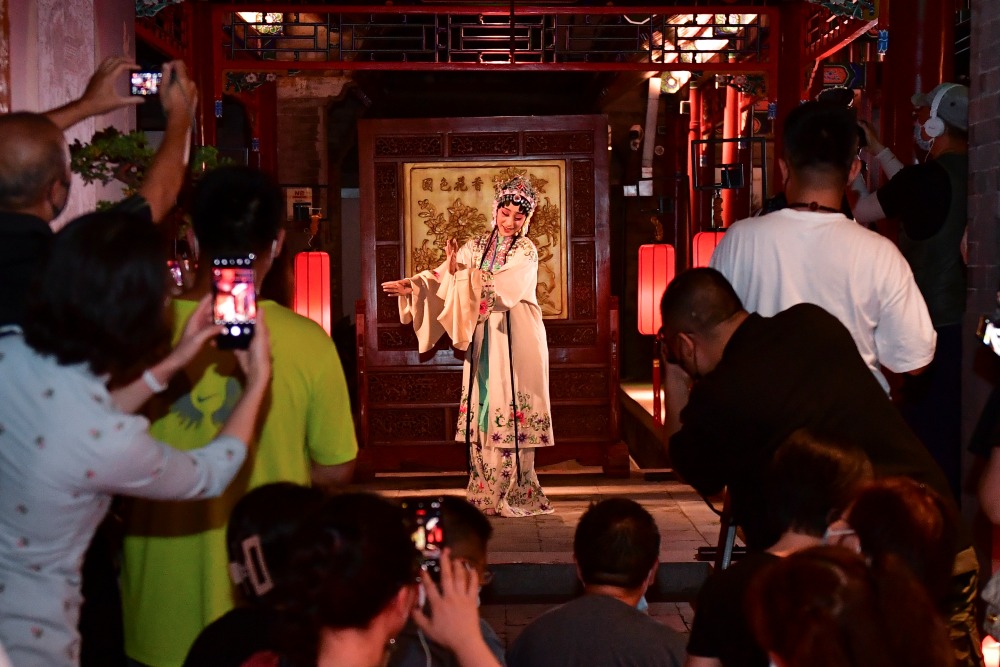 北京正乙祠戲樓內，一名身穿淡色刺繡戲服、頭戴崑曲頭飾的演員在紅色燈籠和木雕屏風前表演；觀眾圍在兩旁觀看，部分人以手機拍攝。