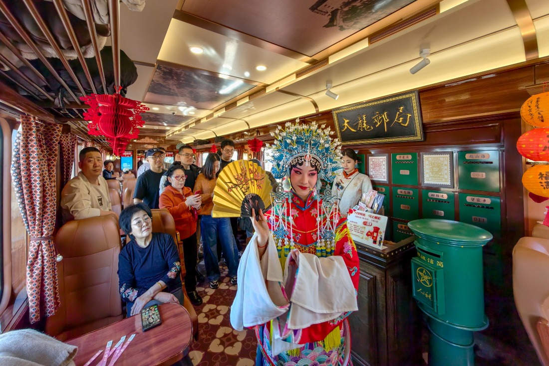 天津「津旅時光號」主題列車車廂內，一名身穿紅色戲服、頭戴藍色頭飾的京劇演員手持摺扇演出；多名乘客坐在座位上或站在車廂通道觀看。