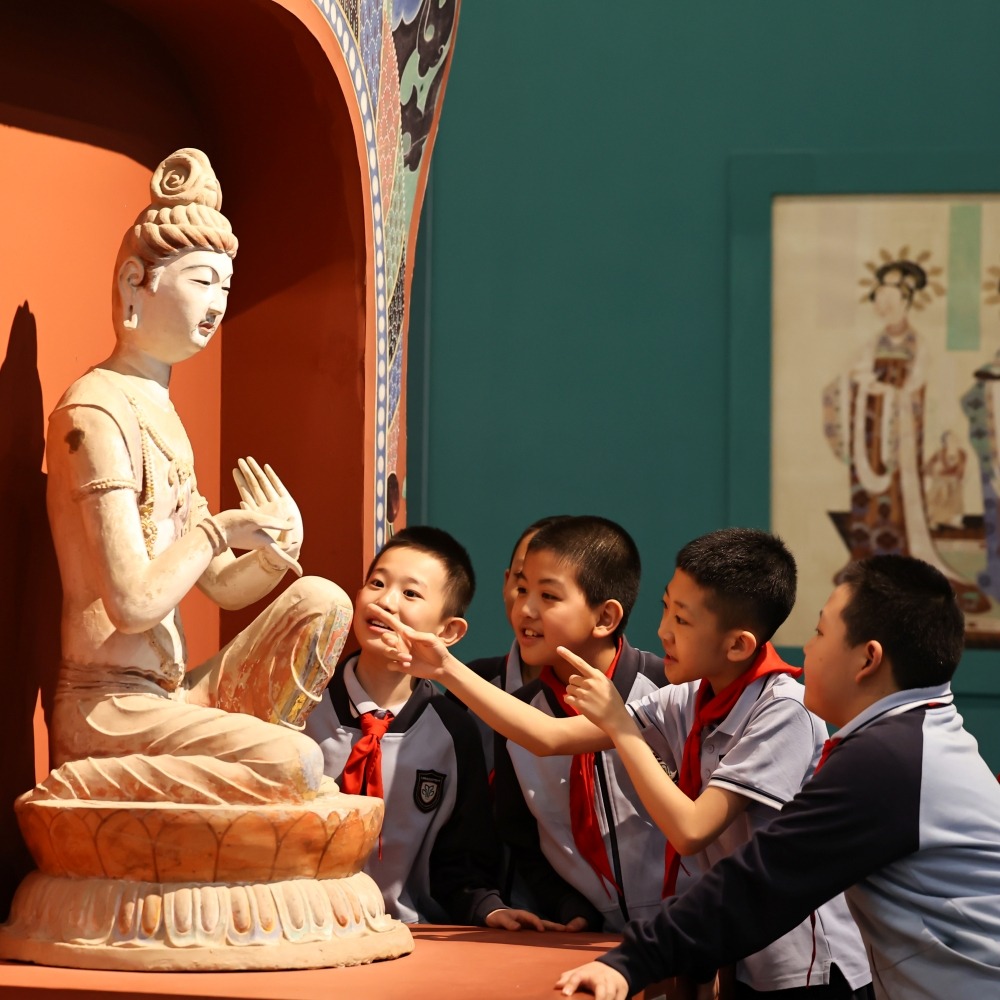  2026年5月18日適逢國際博物館日，小學生到敦煌研究院蘭州敦煌藝術館參觀，在一尊佛像展品前開心觀摩。寒假時，研學學子在敦煌莫高里工匠村參觀。
